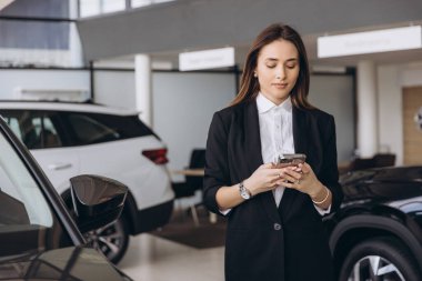 Young saleswoman using mobile phone in a car dealership, choosing a new vehicle for a customer or for herself