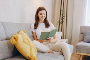 Young woman enjoying a quiet moment of leisure, comfortably seated on her sofa at home, engrossed in reading a captivating book