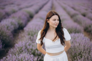 Beautiful woman wearing a flowing white dress, joyfully wandering through vibrant purple rows of a fragrant lavender field in summer