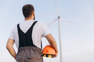 Engineer holding an orange helmet, observing a wind turbine against a backdrop of a clear blue sky, emphasizing renewable energy solutions