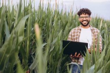 Smiling agronomist holding a laptop while standing confidently in a vibrant corn field, showcasing expertise in sustainable agriculture
