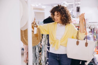 Curly haired woman shopping for summer clothes, holding a beach bag and looking at a straw hat in a fashion retail store