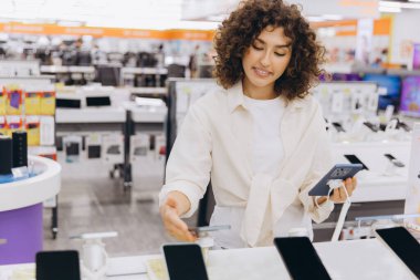 Young woman comparing smartphones in an electronics store, holding one model and looking at another on display, making a purchasing decision