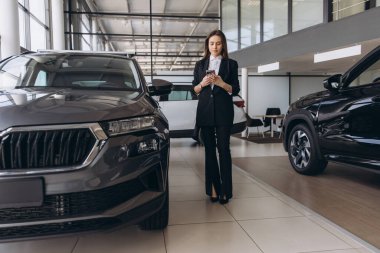 Saleswoman engaging with a mobile phone while positioned next to a sleek new car in a vibrant dealership showroom