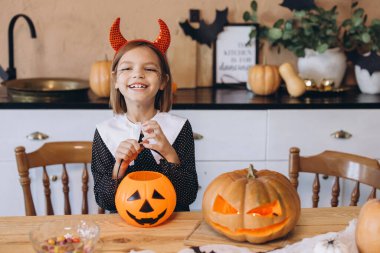 Little girl wearing devil horns headband holding a halloween pumpkin basket with carved pumpkins and candies on a wooden table