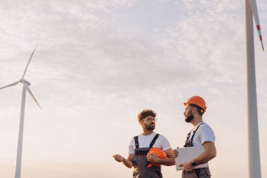 Two engineers discussing maintenance operations while standing next to a wind turbine, collaborating on strategies for sustainable energy solutions