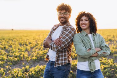 Two smiling agronomists are posing with crossed arms in a soybean field at sunset, showing confidence and teamwork in agriculture