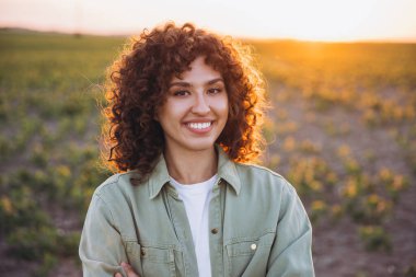 Portrait of a young curly agronomist smiling in a cultivated soybean field at sunset with crossed arms