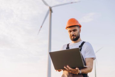 Engineer is holding a laptop and working in a wind turbine farm, contributing to sustainable energy production
