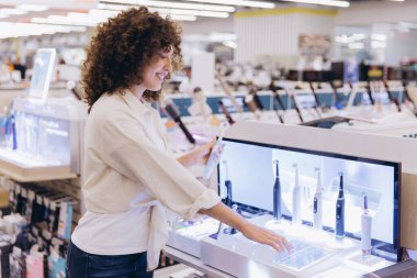 Smiling curly haired woman comparing electric toothbrushes in an electronics store, using a touchscreen to check product information