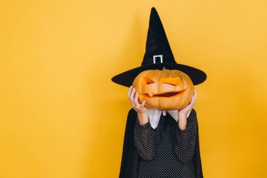 Little girl wearing witch costume hiding her face with halloween pumpkin posing on yellow background