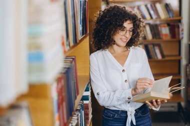 Young curly haired student wearing eyeglasses reading a book standing between bookshelves in a library, enjoying her studying time