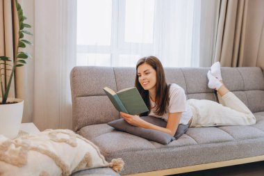 Smiling woman enjoying a relaxing day at home, lying on the sofa and reading a book, creating a cozy and peaceful atmosphere
