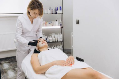 Beautician wearing black gloves applying a white mask on a young woman's face lying on a massage table in a modern beauty salon