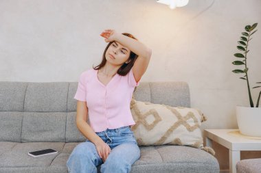 Exhausted young woman touching her forehead while sitting on the sofa at home, suffering from fever or headache