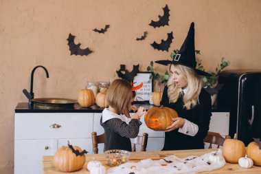 Mom wearing witch costume holding jack o' lantern while daughter carving pumpkin for Halloween party