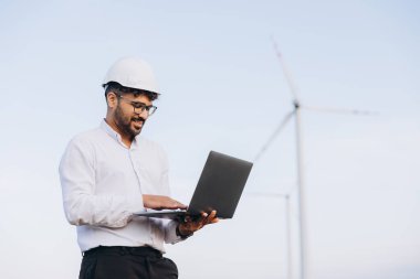 Young engineer working outdoors at a wind turbine power plant, using a laptop to monitor and analyze renewable energy systems