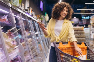 Smiling woman pushing shopping cart and choosing meat from refrigerated section in supermarket