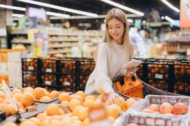 Marketten taze greyfurt seçerken gülümseyen genç bir kadın alışveriş listesini kontrol ediyor ve market deneyiminin tadını çıkarıyor.