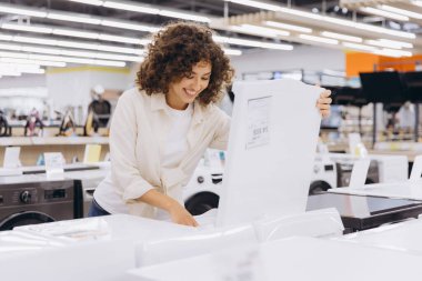 Smiling woman with curly hair browsing and comparing options for a new washing machine in a bustling electronics store