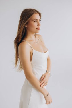 Studio portrait of a young woman with long brown hair wearing a white dress and jewelry, posing against a white background