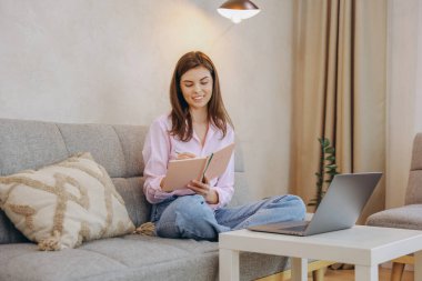 Smiling woman writing on a notebook while sitting on a sofa with a laptop nearby, working from home