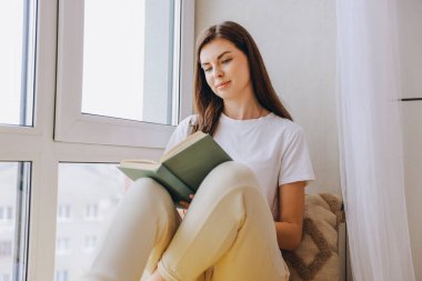 Girl enjoying free time, reading a book while sitting comfortably by the window in her cozy home, basking in natural light