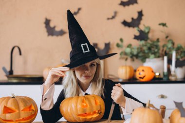 Woman wearing a witch hat joyfully carving Halloween pumpkins in a kitchen adorned with playful paper bats and festive decorations