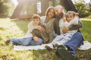 Smiling parents hugging children while sitting on blanket in backyard near modern a frame house