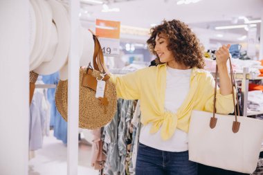 Smiling woman with curly hair enjoying a shopping trip, selecting a stylish round straw bag in a vibrant fashion store
