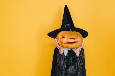 Little witch wearing black hat and dress holding carved pumpkin in front of her face posing on yellow background for Halloween