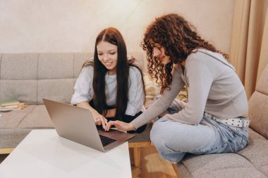 Two teenage girls are using a laptop, enjoying their time together on a comfortable sofa in a cozy home environment
