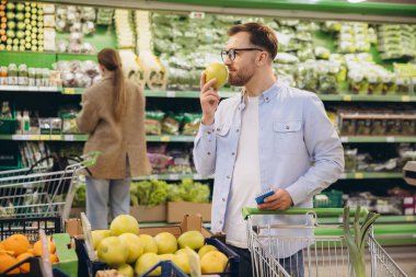 Customer smelling a green apple while holding a shopping list and pushing a cart in a supermarket