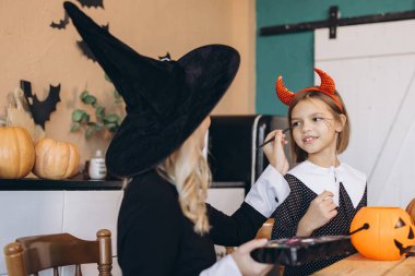 Mother and daughter joyfully applying Halloween makeup while dressed in fun costumes, creating cherished memories at home during autumn festivities