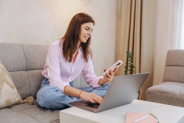 Freelancer managing her business from the comfort of home, using a laptop and smartphone while enjoying a relaxed atmosphere in the living room