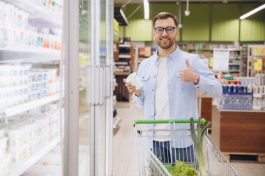 Man doing grocery shopping holding a yogurt and showing thumb up in supermarket