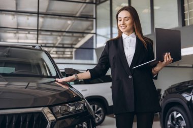 Smiling saleswoman presenting a new car to a customer while holding a laptop in a modern car dealership showroom