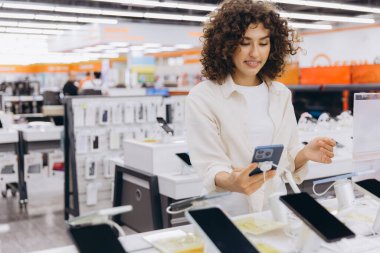 Young woman choosing smartphone in consumer electronics store, comparing different models and features before making purchase