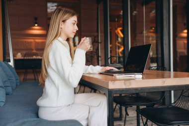 Young woman working remotely on a laptop while enjoying tea in a trendy cafe, embracing a cozy atmosphere and modern lifestyle