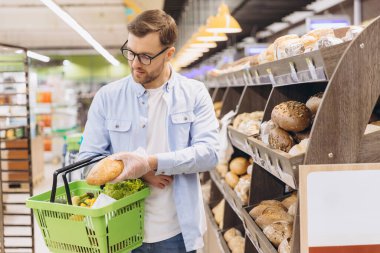 Man choosing bread from shelf in supermarket bakery section, wearing gloves and holding shopping basket