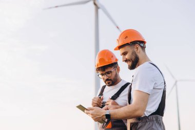 Two engineers collaborating with a tablet beside a wind turbine, discussing advancements in sustainable energy solutions and technologies