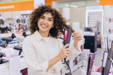 Smiling woman testing new hair straightener in electronics store, choosing best hair care product