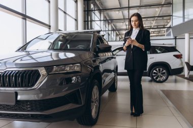 Saleswoman engaging with her smartphone while standing beside a new car in a vibrant car dealership showroom, showcasing modern technology
