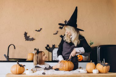 Mom wearing witch costume carving pumpkin with knife while daughter helping her in decorated kitchen
