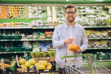 Smiling customer choosing oranges in supermarket with shopping cart