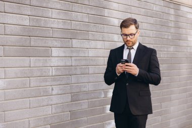 Businessman wearing suit and tie using smartphone standing near gray brick wall