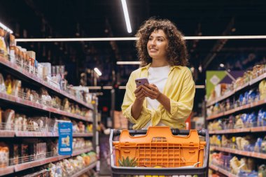 Customer using a smartphone while pushing a shopping cart through a supermarket aisle, selecting fresh groceries and checking items off a list