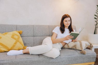 Young woman enjoying her leisure time reading a book while relaxing on a comfortable sofa in her modern living room