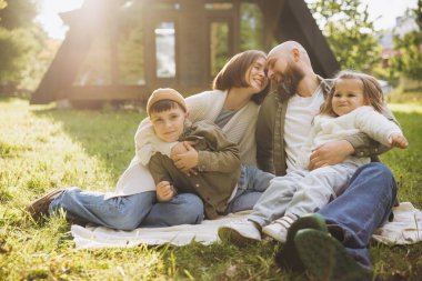 Happy parents hugging their children, sitting on blanket in backyard of their modern a frame house, enjoying sunny day