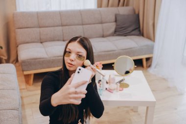 Young woman wearing hydrogel eye patches applying makeup recording video tutorial with smartphone sitting on the floor in the living room